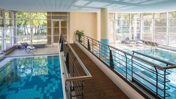 Una piscina interna con un solarium in legno, ringhiere in vetro e poltroncine da relax.