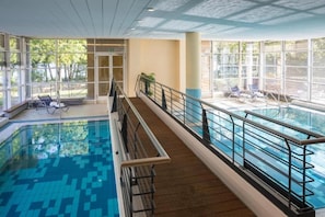 Una piscina interior con cubierta de madera, barandillas de vidrio y sillas de descanso.