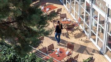 Una terrazza esterna con tavoli e sedie, una persona che cammina e un edificio con finestre vetrate.