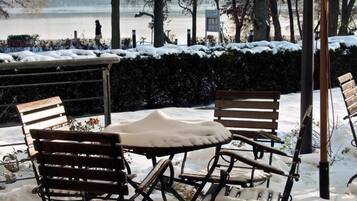 Un área de estar al aire libre con una mesa y cuatro sillas cubiertas de nieve, rodeada de suelo y árboles nevados.