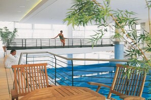 Una piscina interior con un trampolín y una persona saltando. En primer plano, hay sillas de madera y una mesa.