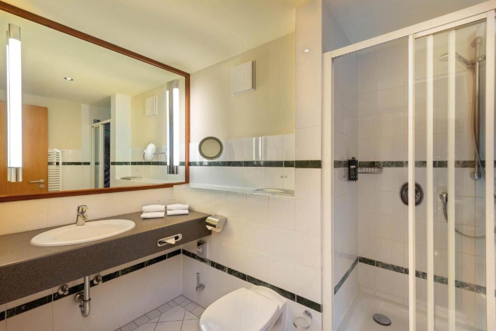Baño de hotel con lavamanos, espejo, toallero y cabina de ducha con mampara de vidrio.