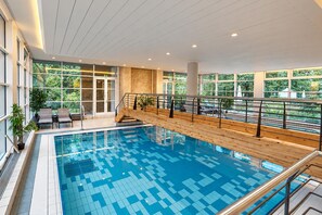 An indoor swimming pool with a tiled floor, a wooden deck with railings, and a view of greenery outside.