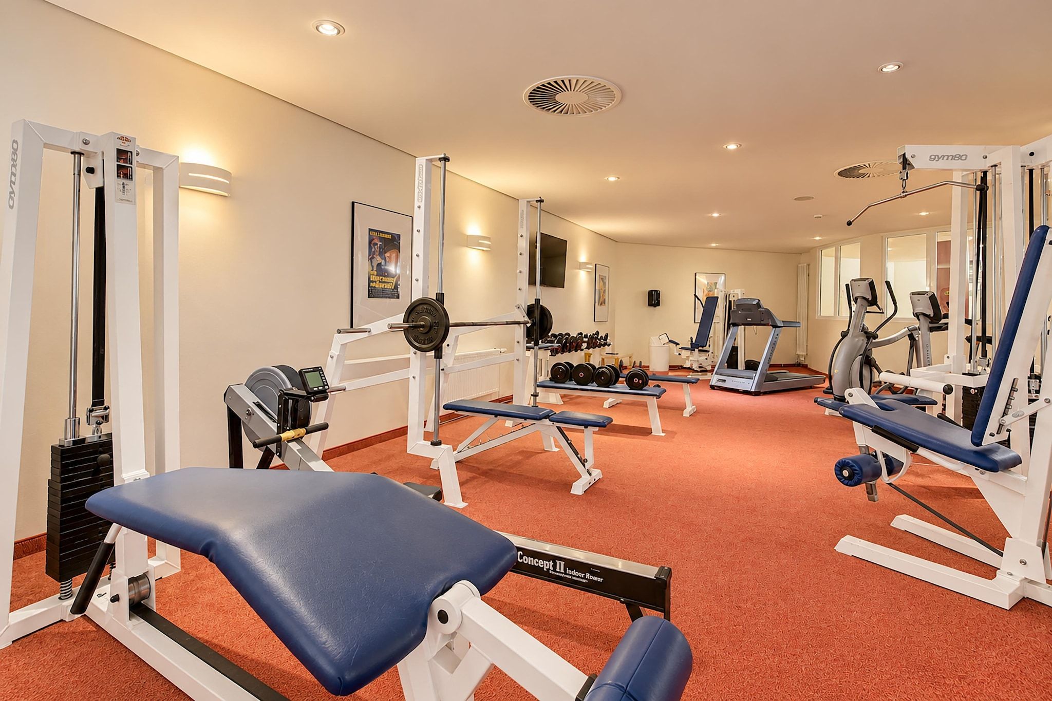 Ein Fitnessstudio mit verschiedenen Trainingsgeräten, darunter eine Hantelbank, Kurzhanteln und ein Laufband.
