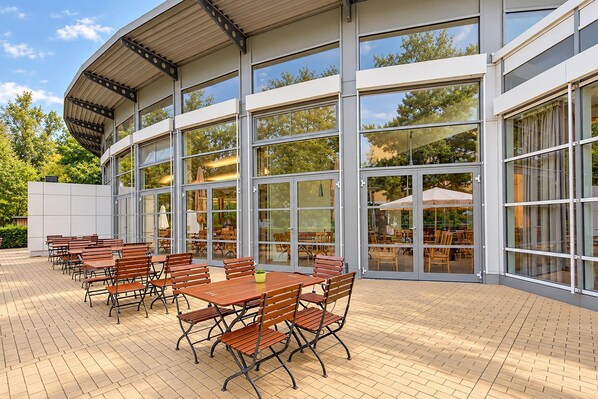 A modern building with large glass windows and a covered outdoor seating area with wooden tables and chairs.