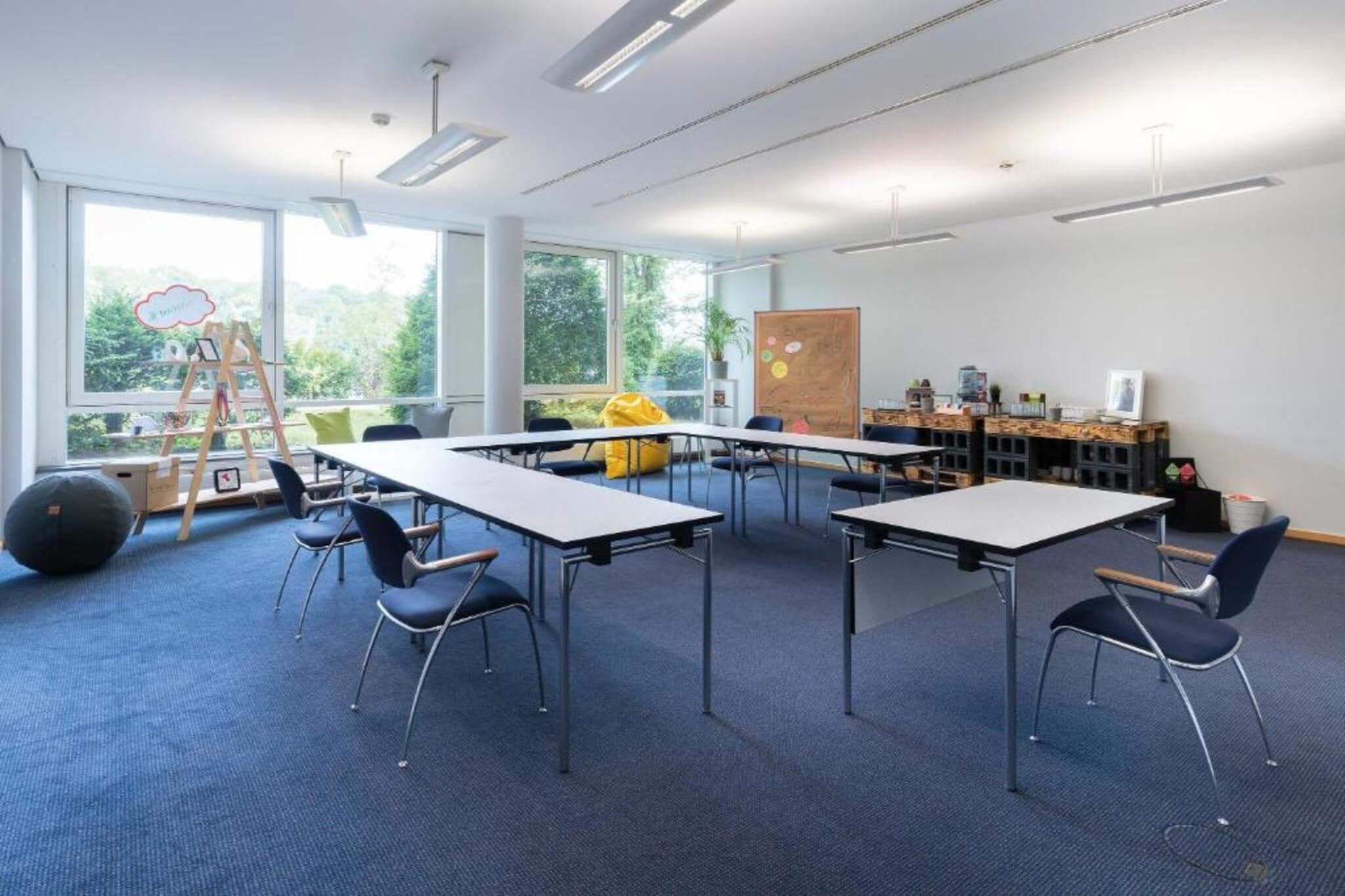 Ein Klassenzimmer mit Tischen und Stühlen, einer Tafel und einem Fenster mit Blick auf Grünflächen.