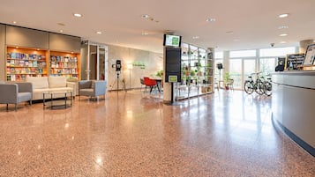 A spacious lobby with a polished floor, seating area, bookshelves, a camera on a tripod, and bicycles in the background.