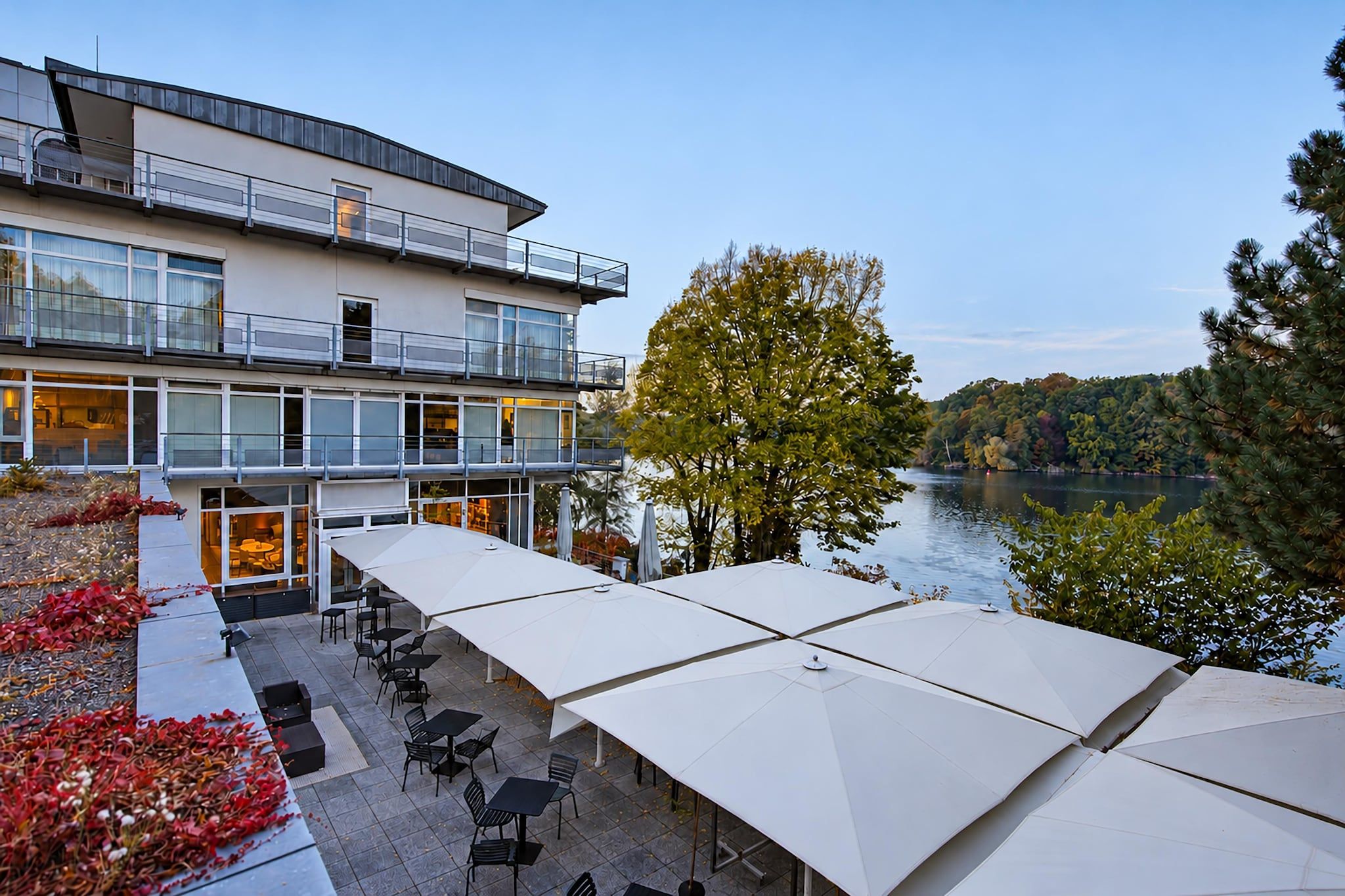 Un edificio moderno con un balcone che si affaccia su un lago, arredi esterni con ombrelloni e fogliame autunnale.