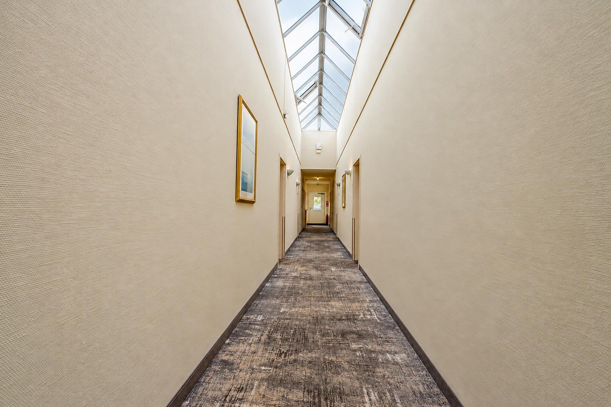 A hotel corridor with a skylight, carpeted floor, and framed artwork on the walls.