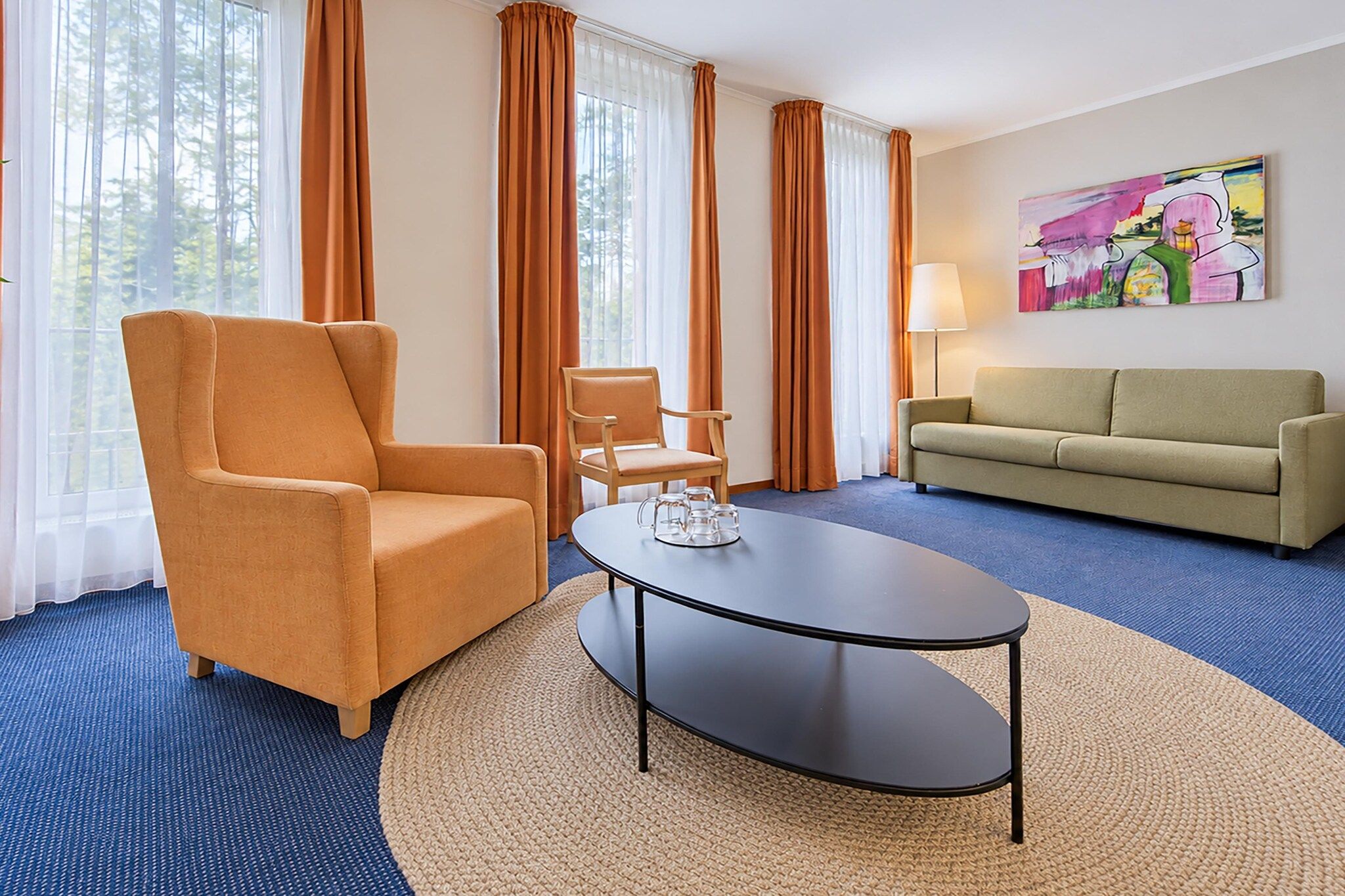 A hotel room with a blue carpet, a round table with a glass centerpiece, a sofa, two armchairs, and a painting on the wall.