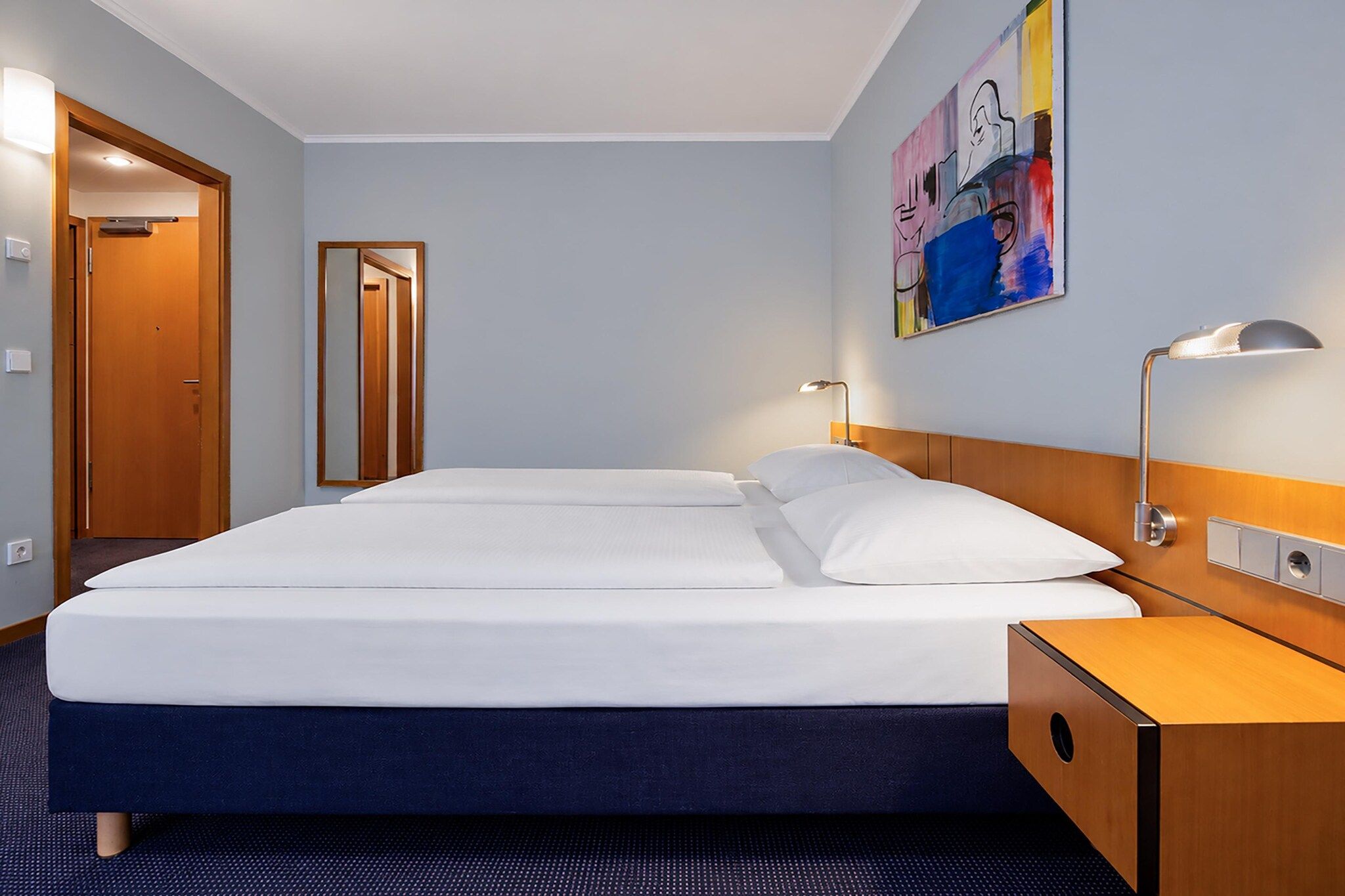 Ein Hotelzimmer mit einem Doppelbett, Nachttischlampen, einem Holznachttisch, einem Spiegel und einem farbenfrohen abstrakten Gemälde an der Wand.