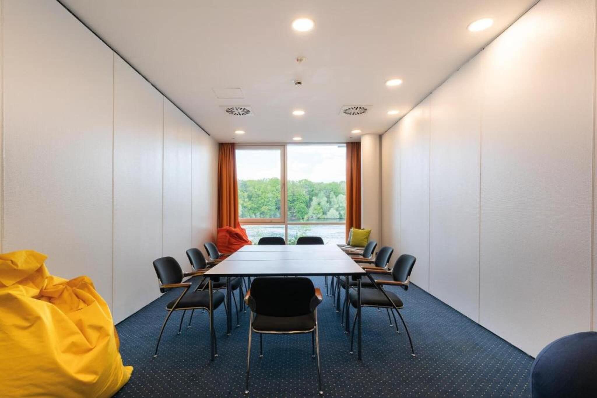 Un salón de conferencias con una larga mesa, sillas y vistas a árboles a través de amplias ventanas.