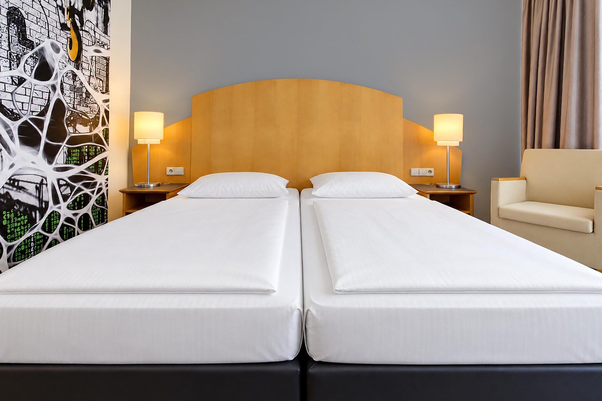 Ein Hotelzimmer mit zwei Einzelbetten, einem Holzkopfteil, Nachttischlampen, einem Sessel und einer Wand mit einer digitalen Kunstausstellung.