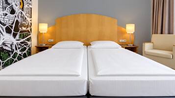 Ein Hotelzimmer mit zwei Einzelbetten, einem Holzkopfteil, Nachttischlampen, einem Sessel und einer Wand mit einer digitalen Kunstausstellung.