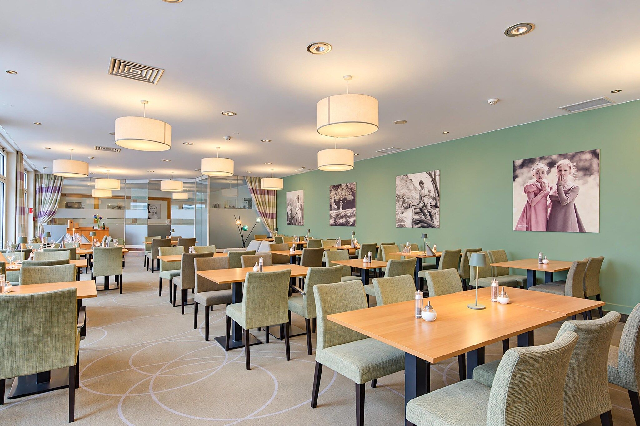 Ein Essbereich eines Hotels mit mehreren Tischen und Stühlen, Deckenleuchten und Wandbildern.