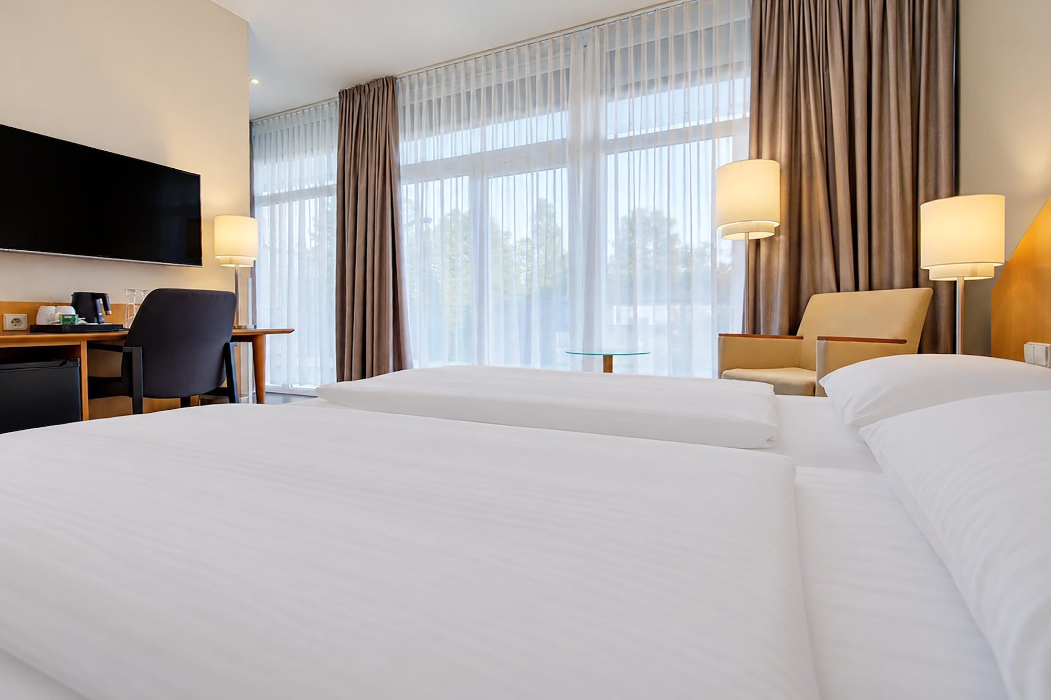 Ein Hotelzimmer mit zwei Betten, einem Schreibtisch, einem Stuhl, einem Fernseher und einem Fenster mit Vorhängen.