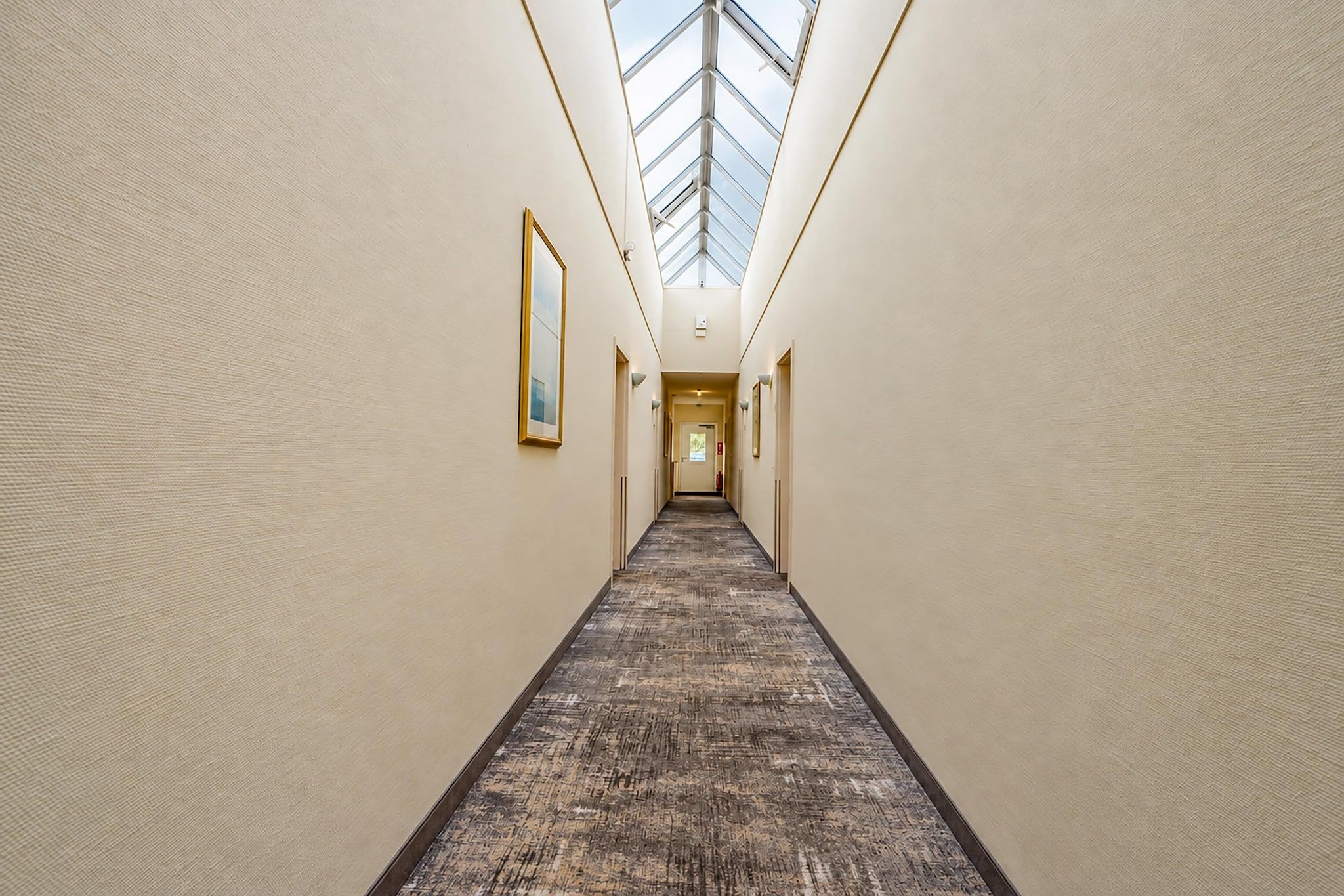 Un couloir d’hôtel avec une verrière, un sol recouvert de moquette et des œuvres d’art encadrées accrochées aux murs.