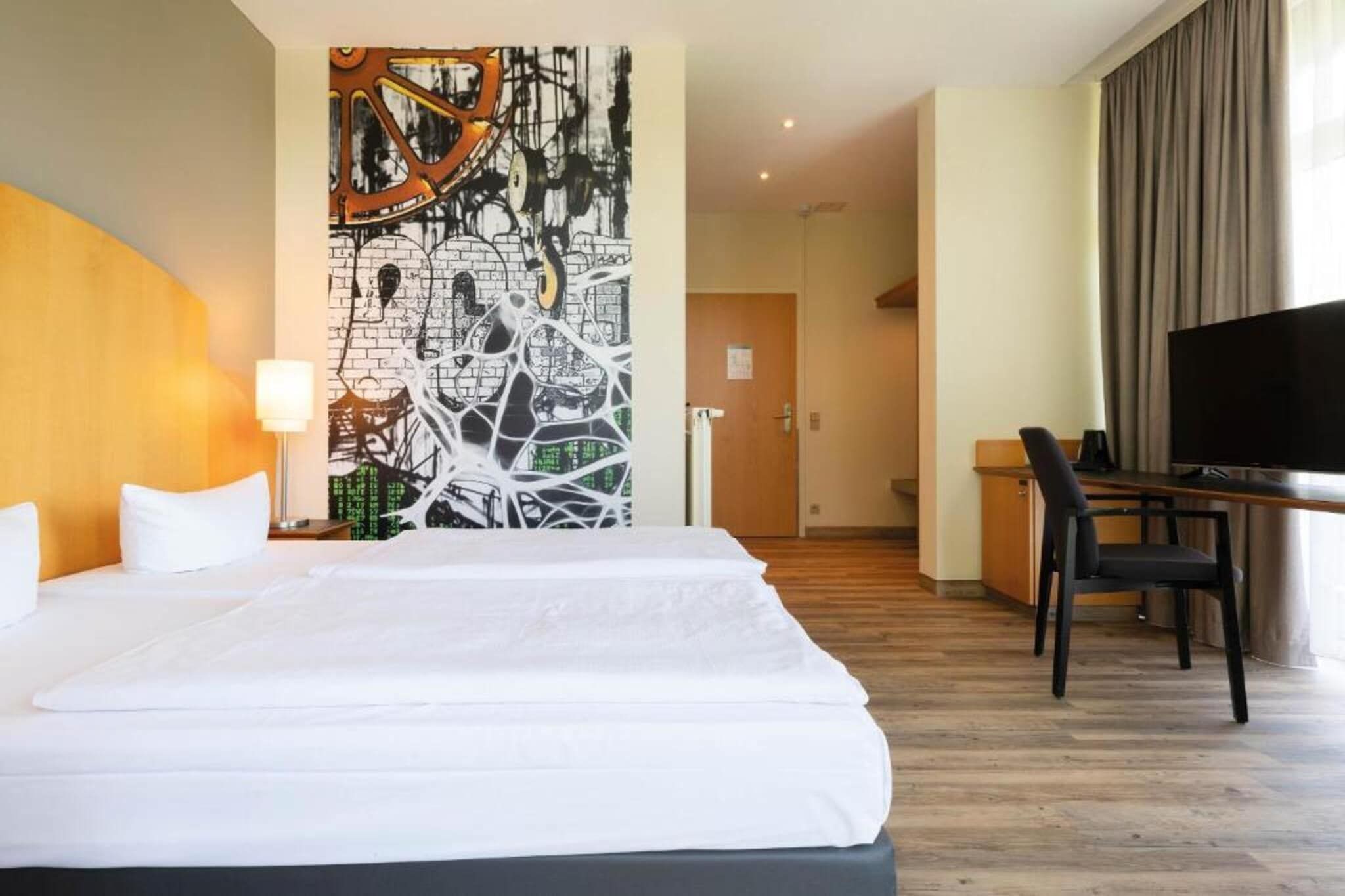 Ett hotellrum med en stor säng, ett skrivbord med en stol, en tv och en vägg prydd med abstrakt konst.