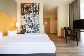 Et hotelværelse med en stor seng, et skrivebord med stol, et fjernsyn og en væg med abstrakt kunst.