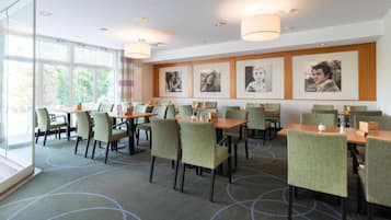 Un comedor de hotel con mesas y sillas, ventanas grandes y cuadros en la pared.