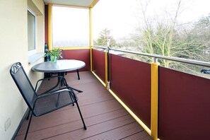 Een balkon met een ronde tafel en twee stoelen, een raam en uitzicht op bomen.