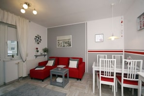 A compact living space with a red sofa, a small coffee table, a dining table with white chairs, and a single pendant light. - Holiday apartment with 1 bedroom (Seebad Heringsdorf)