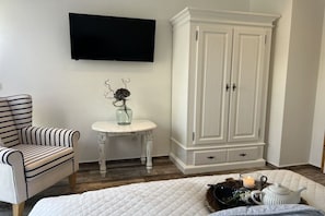 A bedroom with a bed, a television mounted on the wall, a striped chair, a small table with a vase, and a white wardrobe. - Apartments Ostseesonne (Sellin\/Moritzdorf auf Rügen)