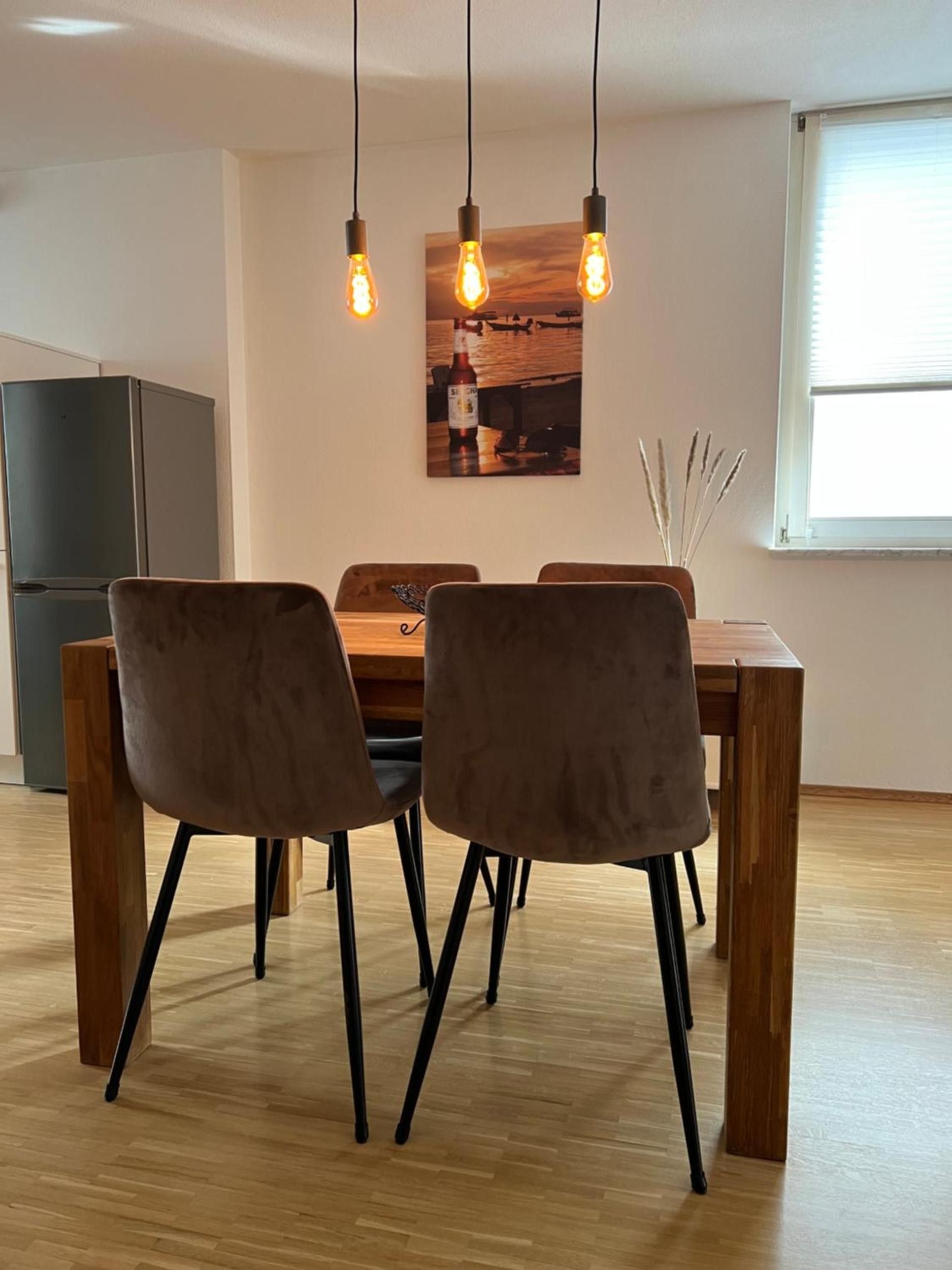 Un comedor con una mesa de madera y cuatro sillas, una lámpara colgante y un refrigerador al fondo.