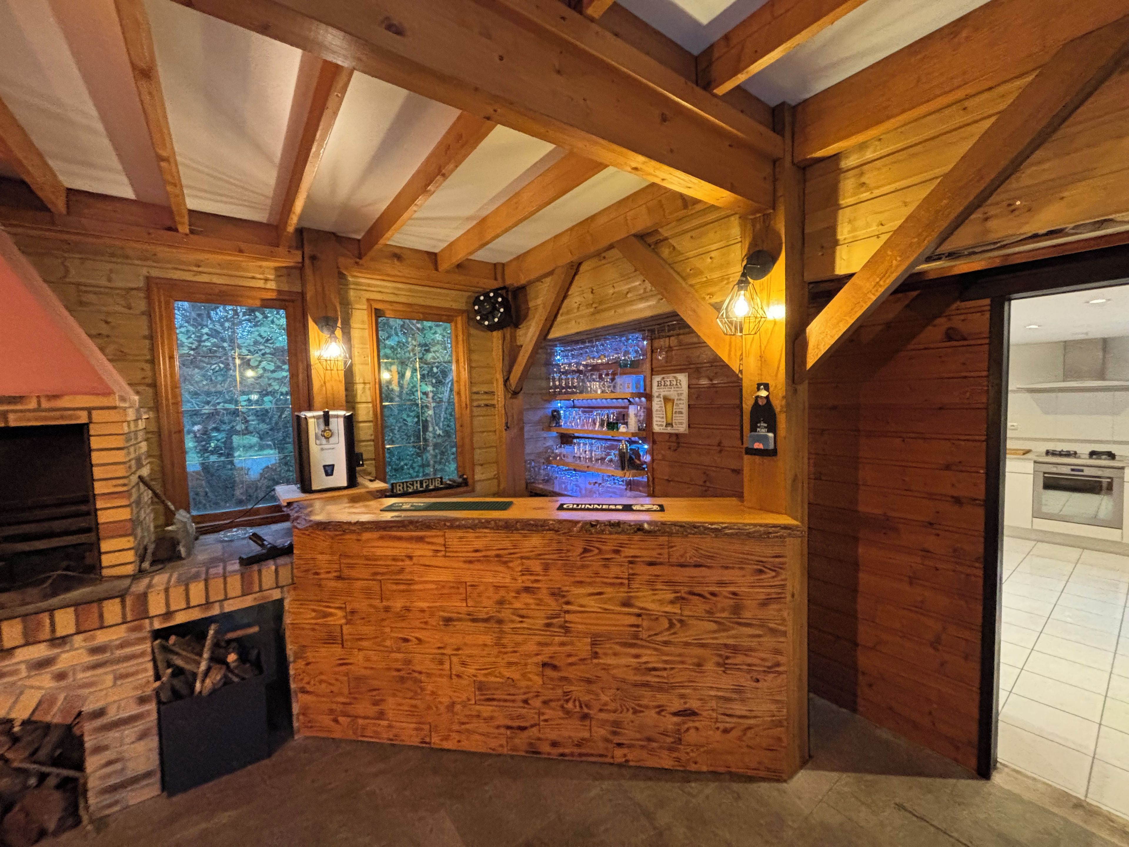 Un coin bar rustique en bois, avec une cheminée en briques, un comptoir et une vue sur la forêt par les fenêtres.