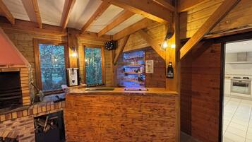Un coin bar rustique en bois, avec une cheminée en briques, un comptoir et une vue sur la forêt par les fenêtres.