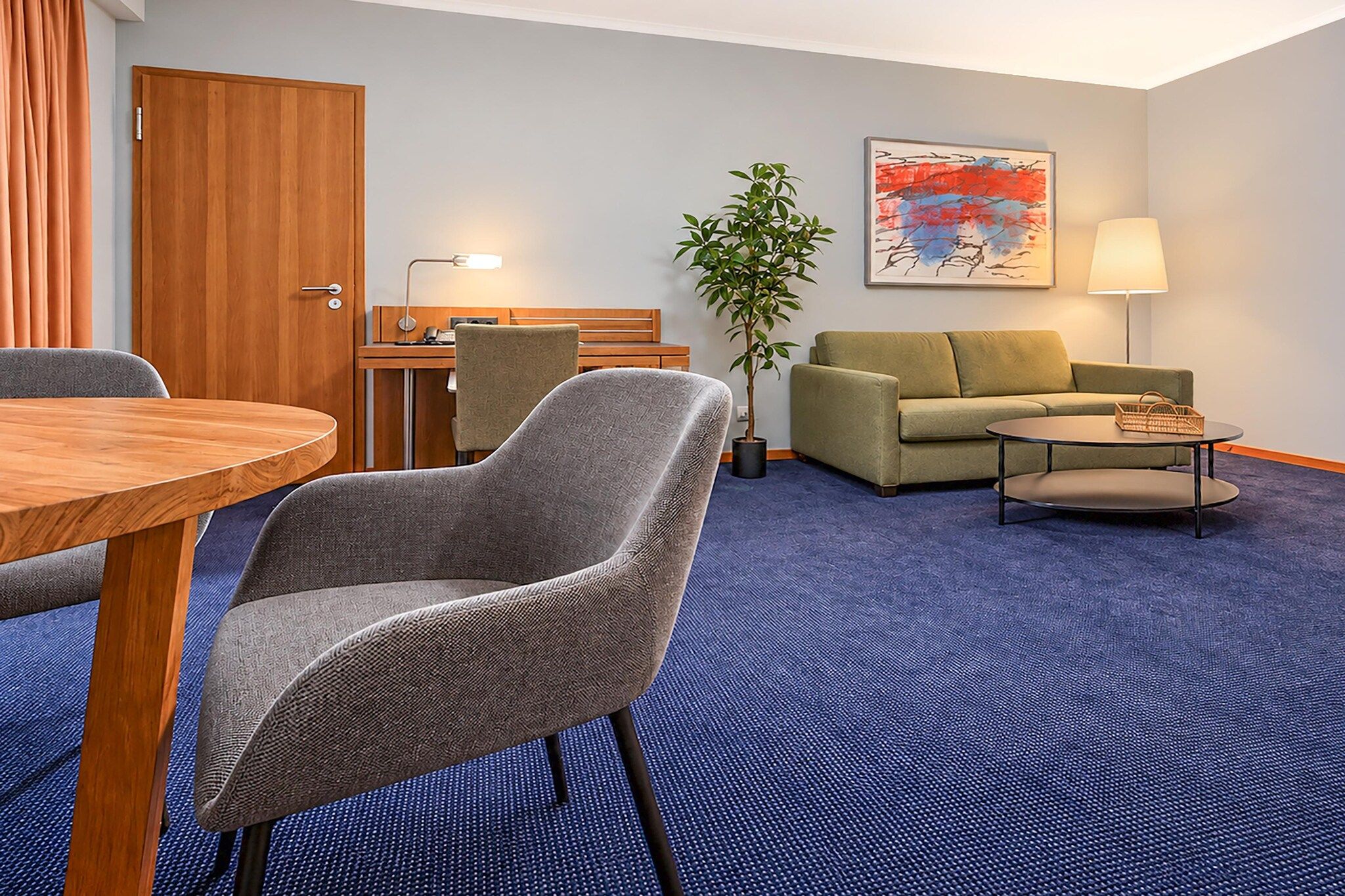 Ein Hotelzimmer mit Esstisch, zwei Sesseln, einer Couch, einem kleinen Couchtisch, einer Lampe, einer Pflanze und einem Bild an der Wand.