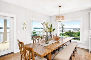 A dining area with a wooden table, chairs, a bench, and a view of a body of water through large windows. - Laguna Matata by Kaiser | Waterfront Little Lagoon | Heated Pool | Near Beach (Gulf Shores)