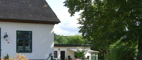 A white building with a thatched roof, a small garden with flowers, and a tree in the background.