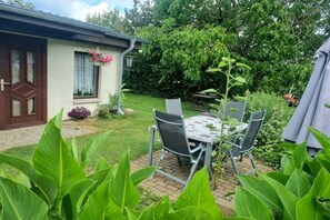 A patio with a table and chairs, a house with a door and window, and a garden with plants. - Bungalow with 2 bedrooms (Großwoltersdorf)