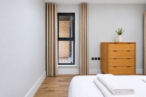 A bedroom with a wooden dresser, a bed with white linens, and a view through a window with curtains. - City Court - Stylish 1BR home in the City (London)