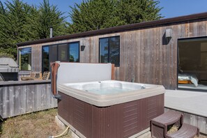 A wooden cabin with a hot tub, a barbecue grill, and a large window. - Lotus Tide | Hot Tub & Golf Course Views (The Sea Ranch)