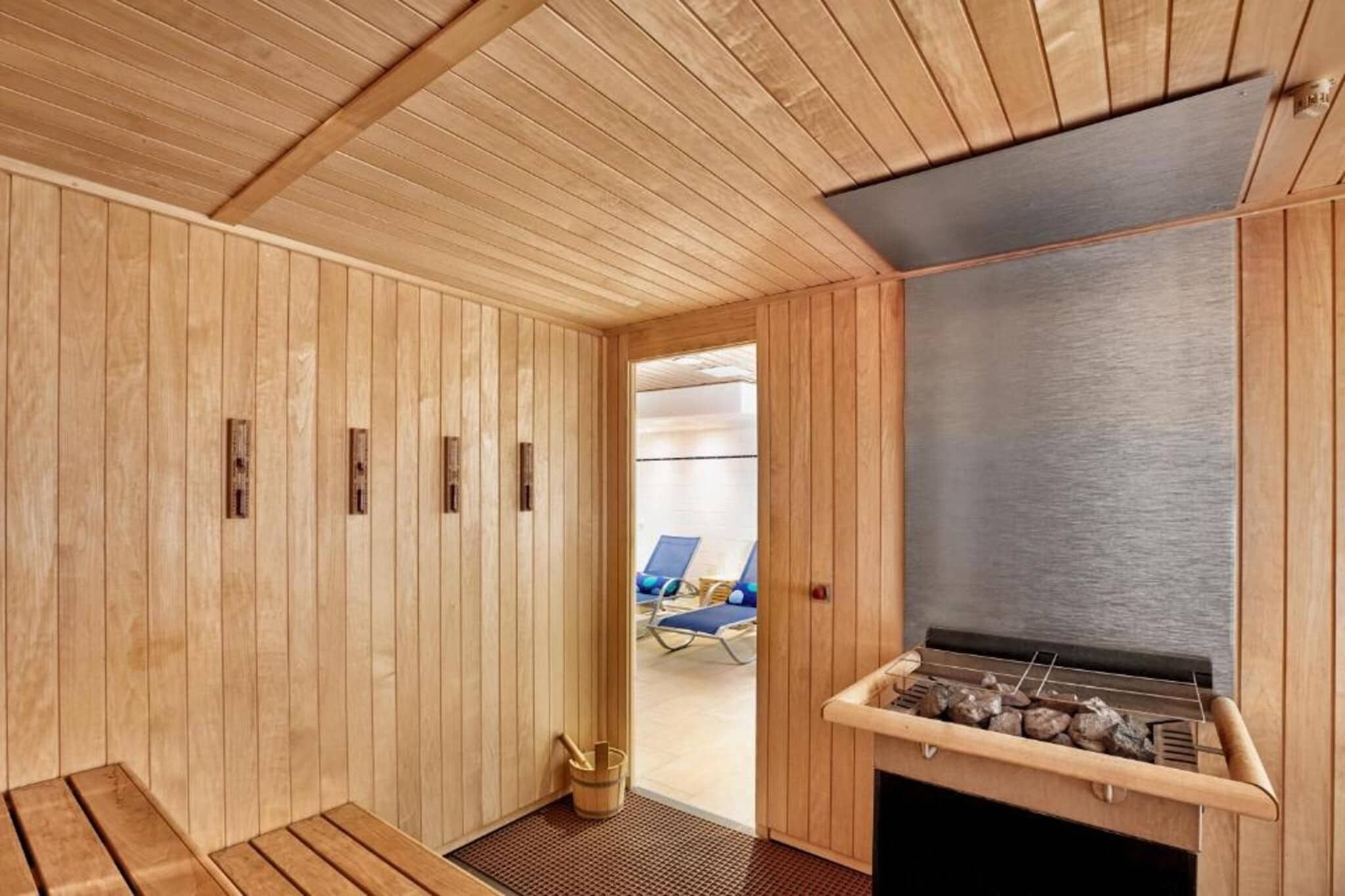 Ein Saunaraum mit Holzwänden, einer Sitzbank, einer Saunaheizung und Blick auf einen Poolbereich mit Liegestühlen.