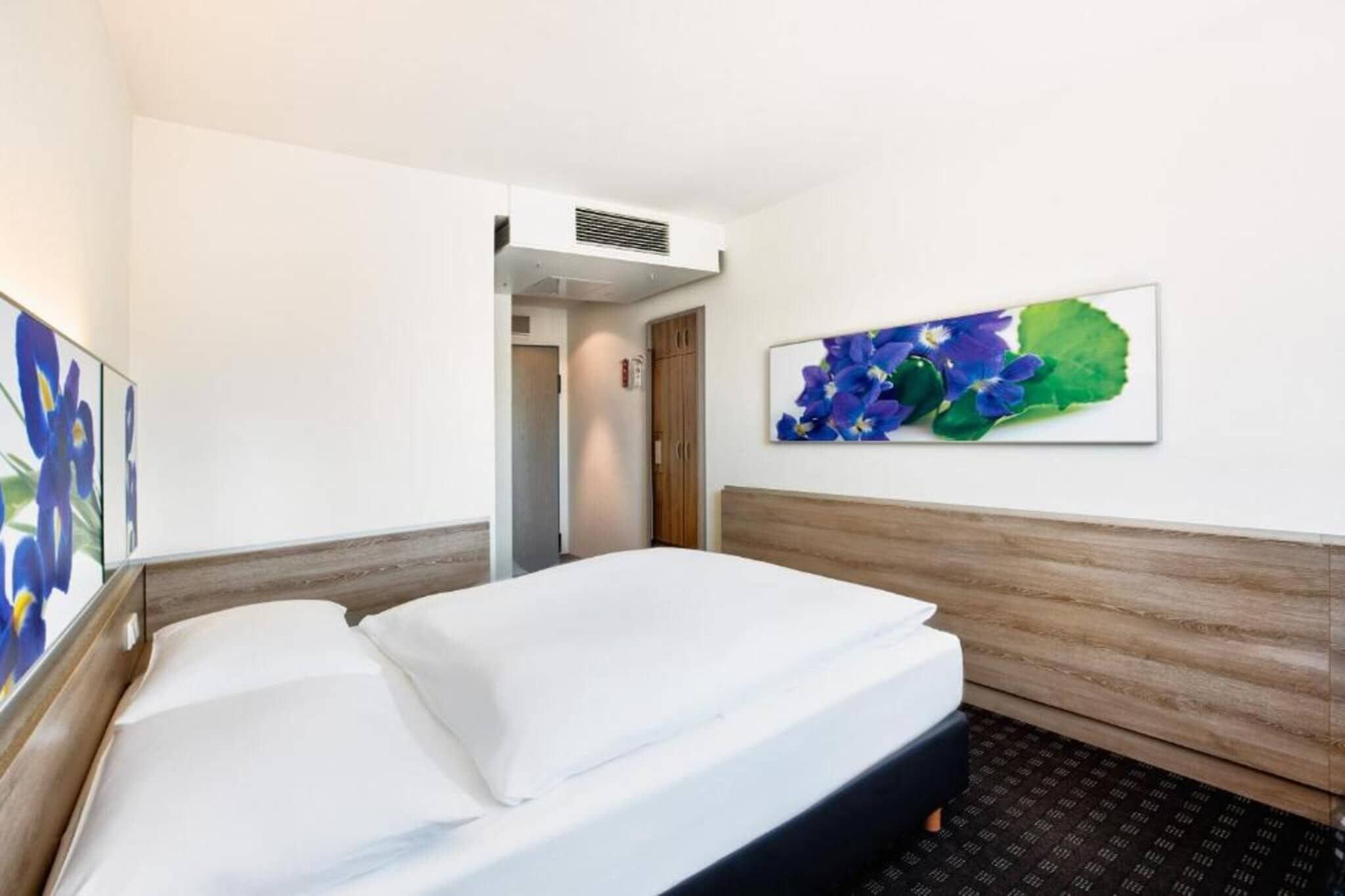 A hotel room with a bed, a door, a wall-mounted air vent, and a framed floral artwork.