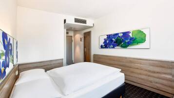 A hotel room with a bed, a door, a wall-mounted air vent, and a framed floral artwork.