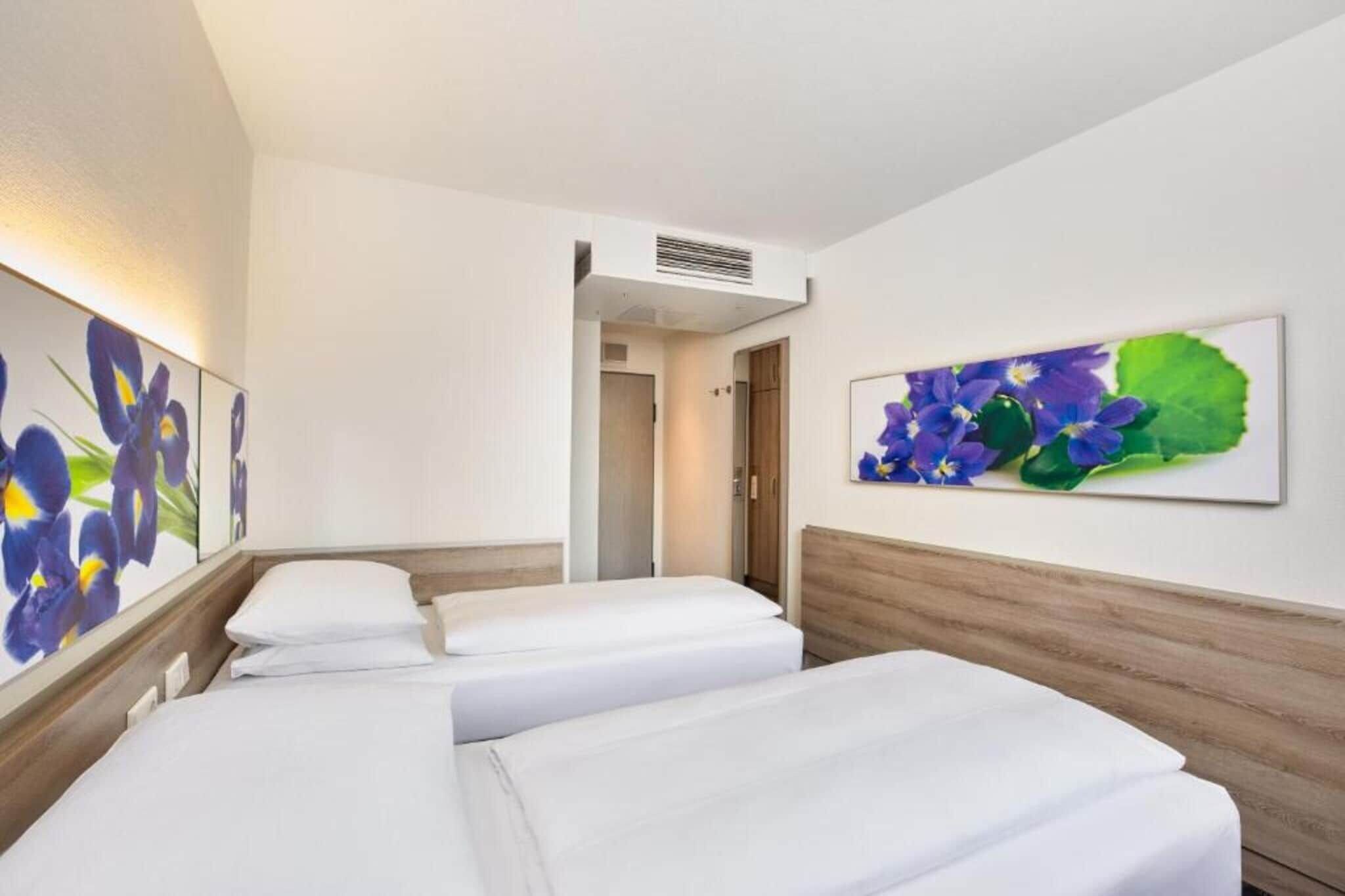 A hotel room with three single beds, a wooden headboard, a wall-mounted air conditioning unit, and two framed floral artworks.