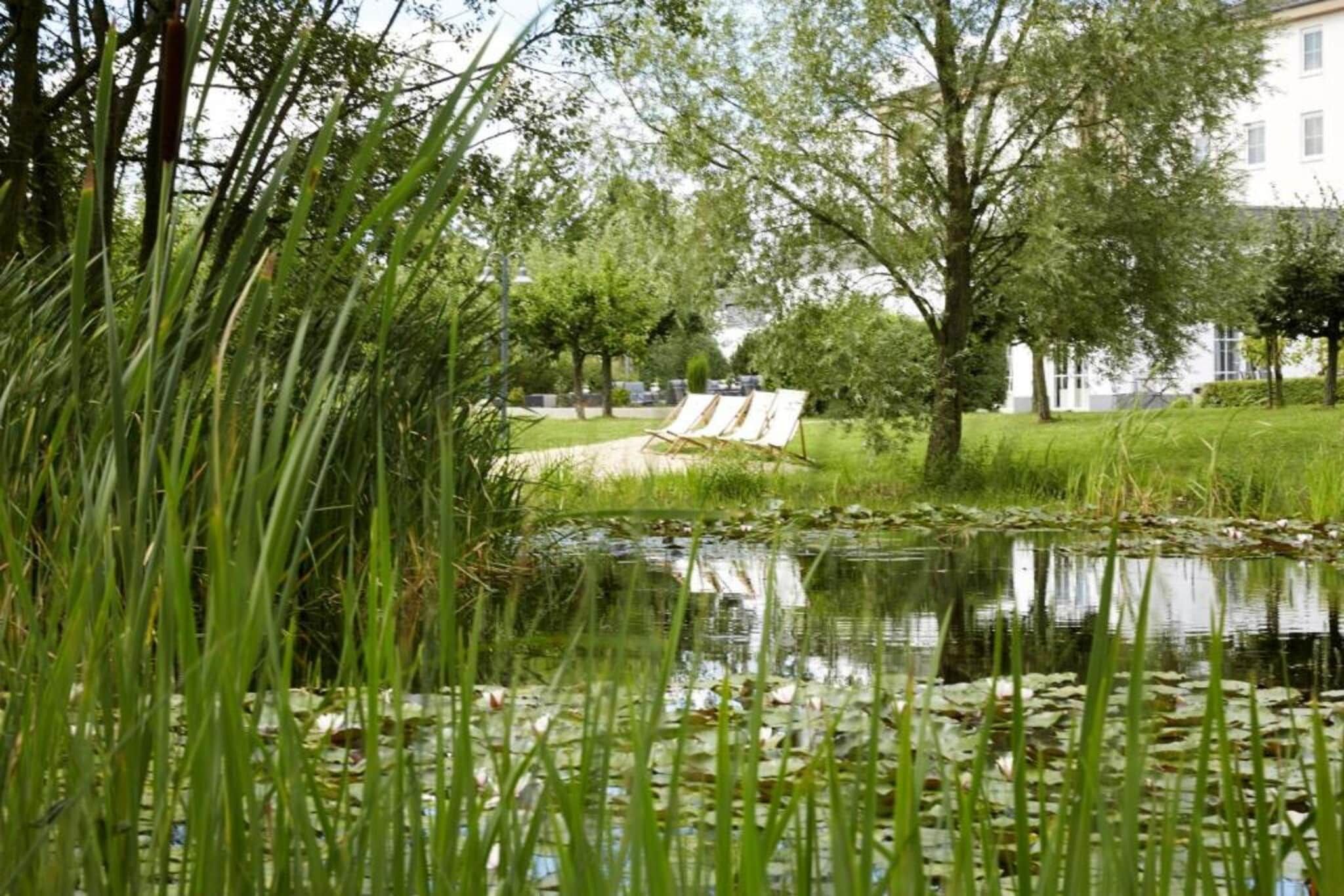 Un’area esterna tranquilla con uno stagno, circondata da vegetazione lussureggiante e alberi. Sono presenti sedie a sdraio e un edificio sullo sfondo.