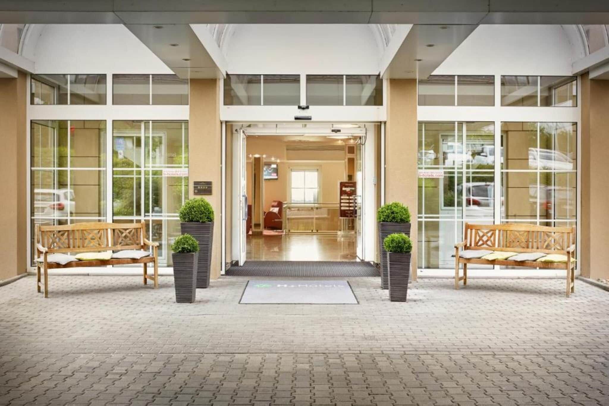 Entrada de hotel con puertas dobles, dos bancos y plantas en macetas.
