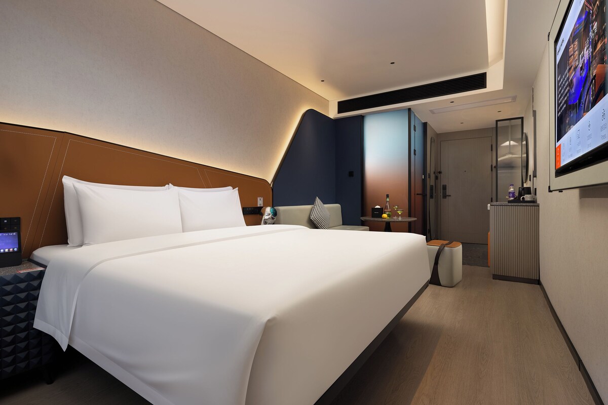 Habitación de hotel moderna con una cama grande, un escritorio, una silla, una pequeña mesa con una bebida y un televisor en la pared.
