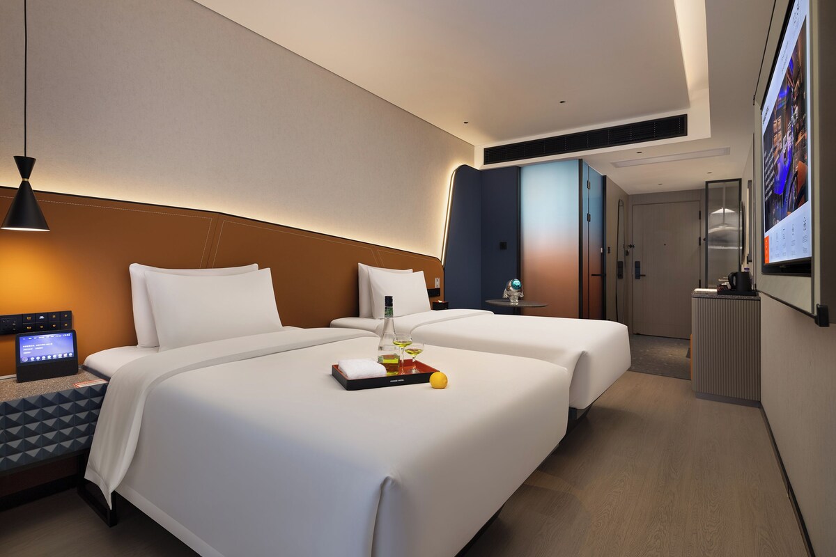 Habitación de hotel con cama, mesita de noche, lámpara y una pantalla digital mostrando información.