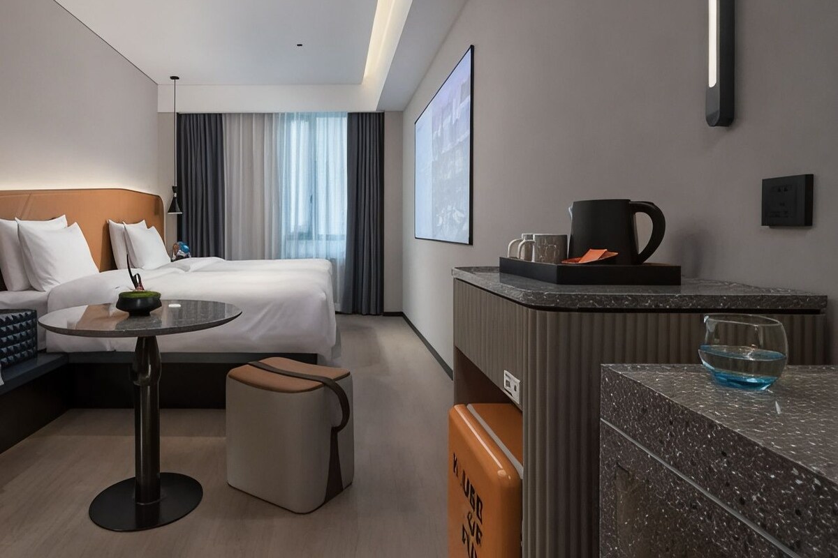 Habitación de hotel con cama, una mesita, zona de estar, mostrador con cafetera y un ventanal con cortinas.