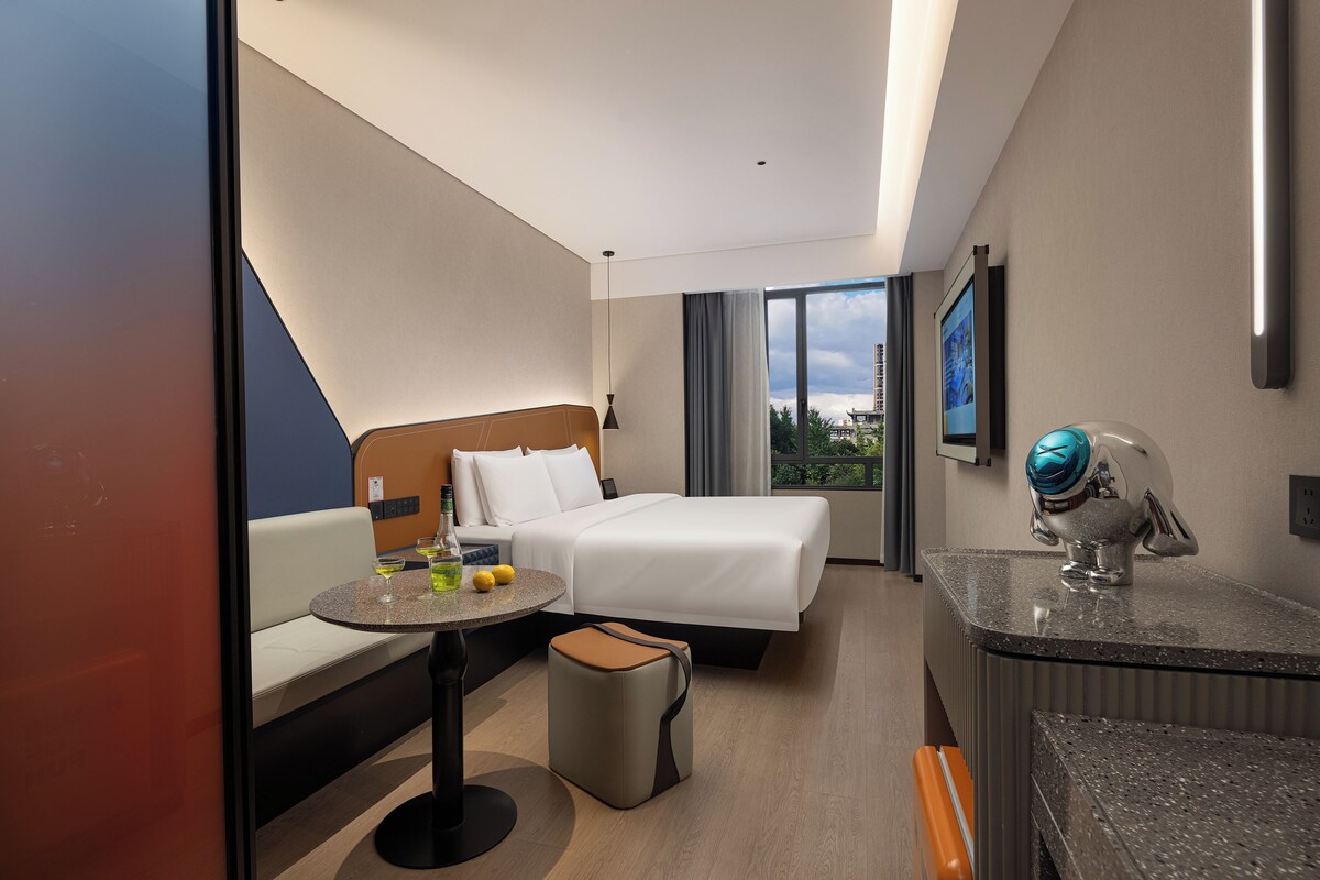 Habitación de hotel con cama, sofá, una mesita con una botella y vasos, un cesto de basura y un baño con espejo y perchero para toallas.