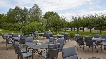 Een buiten eethoek met meerdere tafels en stoelen, omringd door groen en bomen.