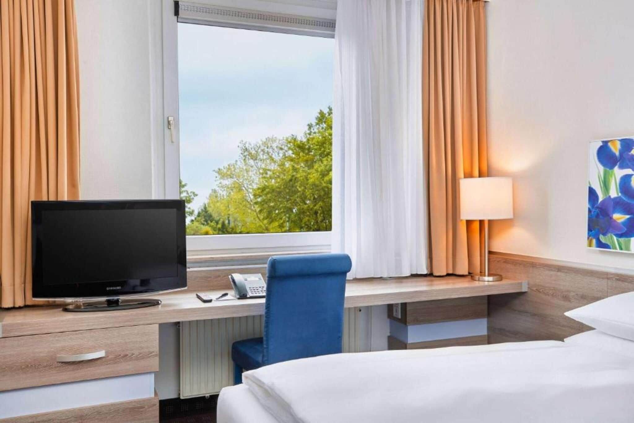 Een hotelkamer met een bed, een bureau met een computer, een stoel, een lamp en een raam met gordijnen.