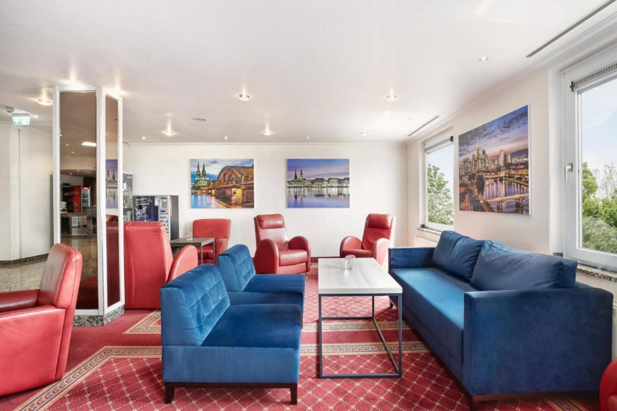Un hall de hotel con sofás rojos y azules, una mesa de centro y grandes ventanales con vistas a la ciudad.