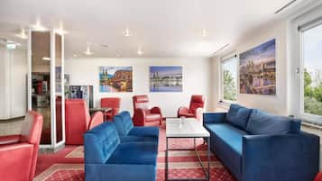 Un hall de hotel con sofás rojos y azules, una mesa de centro y grandes ventanales con vistas a la ciudad.